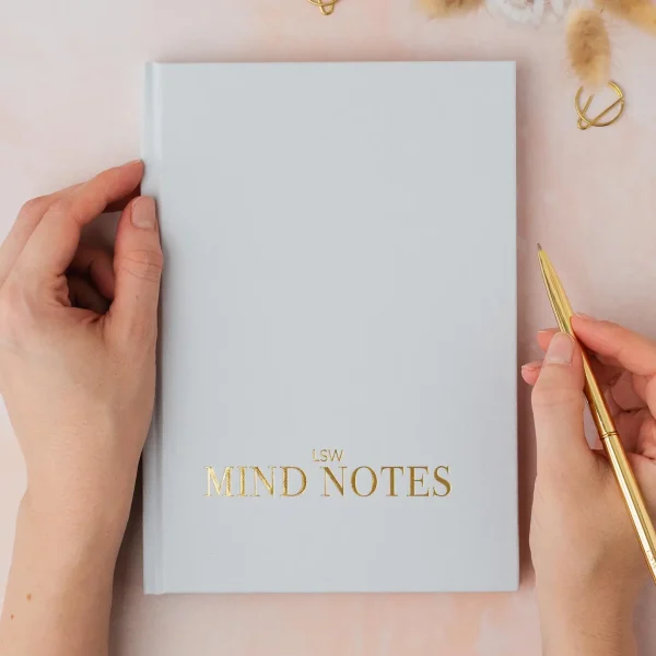 Hands holding a grey LSW Mind Notes journal and a gold pen