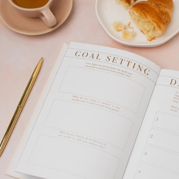 Morning Notes journal open at the goal setting page beside a croissant and coffee